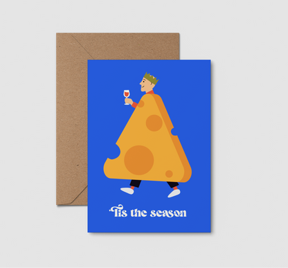A blue greeting card with an illustration of a man in a giant cheese slice costume holding a wine glass, with the text 'Tis the season' at the bottom, shown with a brown kraft envelope. 
