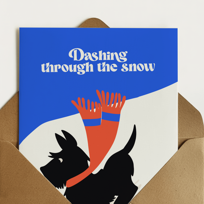 A6 greeting card with a brown kraft envelope, featuring a blue and white design with the text 'Dashing through the snow' above a black dog reaching up with red and blue striped scarf details.