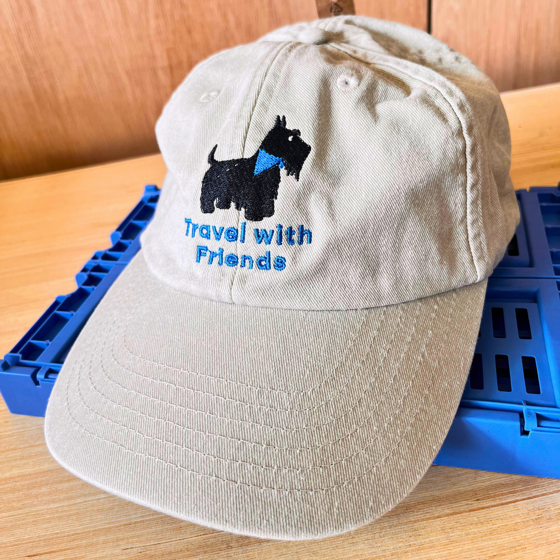 A light beige baseball cap with an embroidered black Scottie dog wearing a blue bandana and the blue text 'Travel with Friends' on the front. Fashion caps.