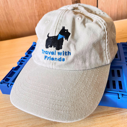 A light beige baseball cap with an embroidered black Scottie dog wearing a blue bandana and the blue text 'Travel with Friends' on the front. Fashion caps.