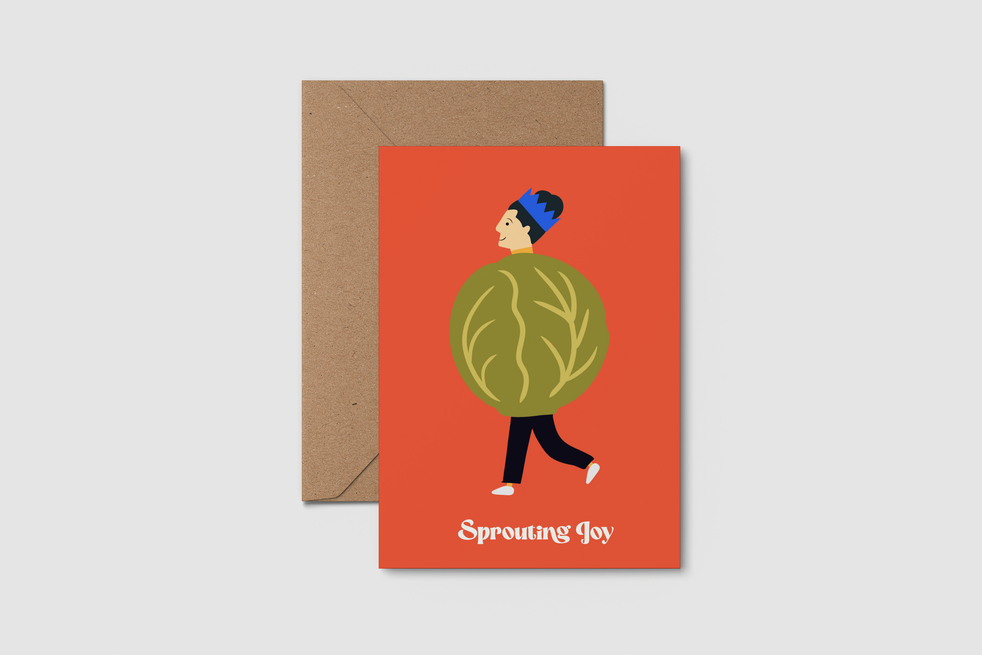 A greeting card with a brown kraft envelope, featuring a festive illustration of a person wearing a brussel sprout against a red-orange background, with the text 'Sprouting Joy' at the bottom.