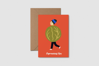 A greeting card with a brown kraft envelope, featuring a festive illustration of a person wearing a brussel sprout against a red-orange background, with the text 'Sprouting Joy' at the bottom.