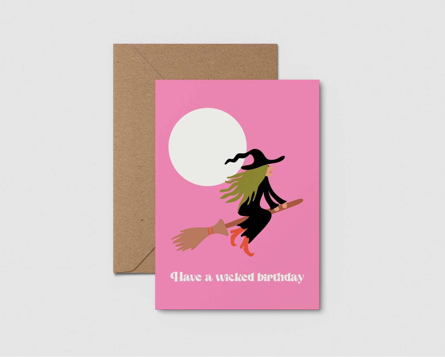 A pink greeting card with a brown kraft envelope, featuring a stylized witch riding a broomstick in front of a white moon and the text 'Have a wicked birthday'.