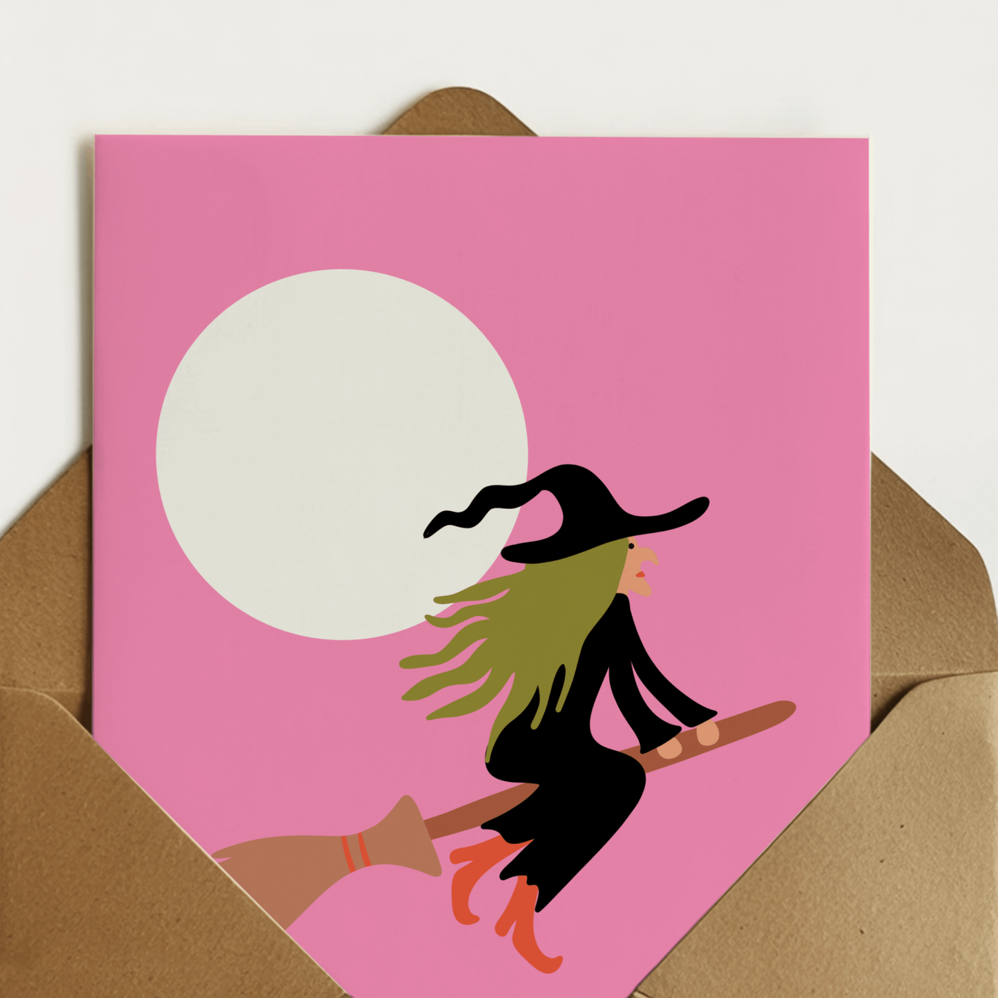 A6 greeting card with a pink background, featuring a witch riding a broomstick in front of a large white moon. A card for spooky season or your neighbourhood witch