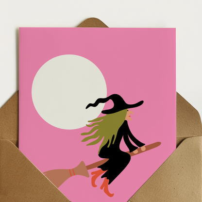 A6 greeting card with a pink background, featuring a witch riding a broomstick in front of a large white moon. A card for spooky season or your neighbourhood witch