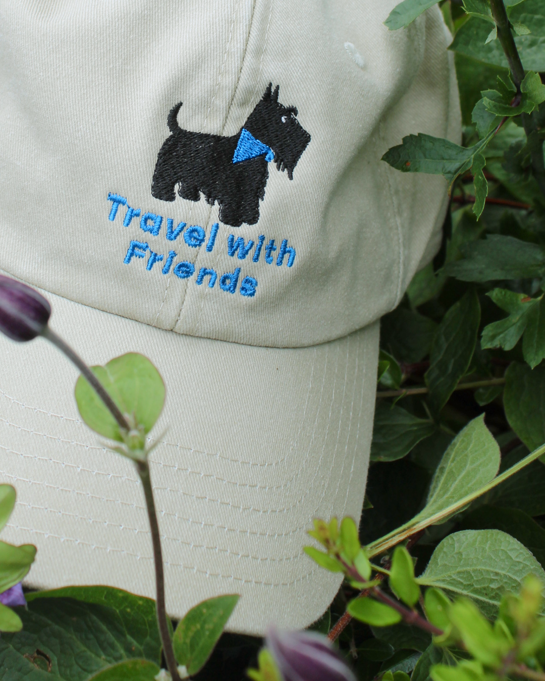 A close-up of a light beige baseball cap with an embroidered black Scottie dog wearing a blue bandana and the phrase 'Travel with Friends' in blue. Cute fashion caps on trend for Autumn.